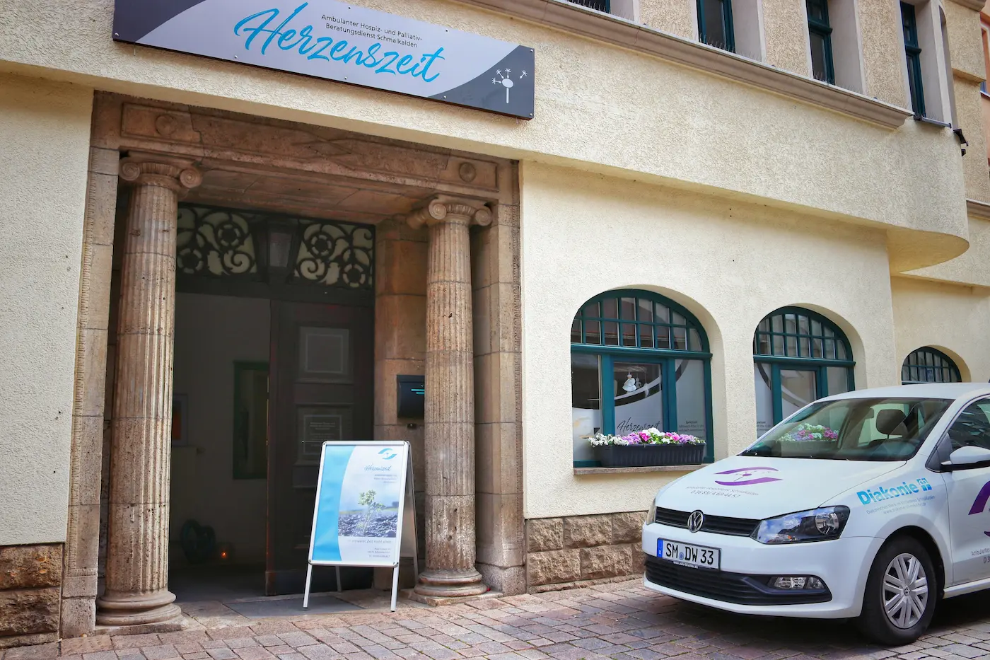 Eingang des Hospizdienstes Herzenszeit Schmalkalden mit Aufsteller, Blumen im Fenster und weißem Dienstfahrzeug davor.
