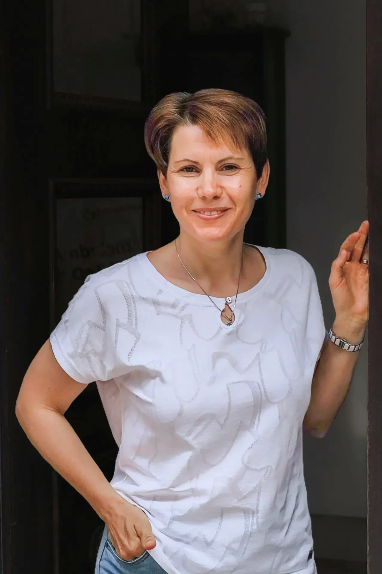 Frau mit kurzem Haar trägt weißes Oberteil und Jeans, steht lächelnd in einer Türöffnung mit einer Hand am Türrahmen.